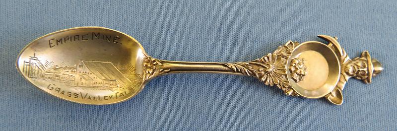 Souvenir Mining Spoon Empire Mine.JPG - SOUVENIR MINING SPOON EMPIRE MINE GRASS VALLEY CA - Sterling silver souvenir spoon,  features  handle with figural miner behind pan with gold nuggets and pick and shovel, flower with deep relief decoration down the handle, bowl has engraved image of mining scene, marking  is EMPIRE MINE, GRASS VALLEY, CAL., measures 4" in length , reverse marked STERLING with hallmark  of H on a pennant for Mechanics Sterling Company, which was subsidiary of Watson Newell Co. [The city of Grass Valley is the largest city in the western region of Nevada County, California.  Situated at roughly 2,500 feet elevation in the western foothills of the Sierra Nevada mountain range, this historic northern Gold Country city is 57 miles north by northeast  from the state capitol in Sacramento. Grass Valley, which was originally known as Boston Ravine and later officially named Centerville, dates from the California Gold Rush, as does nearby Nevada City. When a post office was established in 1851, it was renamed Grass Valley the following year.   The town incorporated in 1860. Grass Valley is the location of the Empire Mine and North Star Mine, two of the oldest, largest, deepest, longest and richest gold mines in California.  Many of those who came to settle in Grass Valley were tin miners from Cornwall, England. They were attracted to the California gold fields because the same skills needed for deep tin mining were needed for hard rock (deep) gold mining. Many of them specialized in pumping the water out of very deep mining shafts.  George Roberts identified the Ophir Hill vein, which would eventually become the Empire Mine, which along with the Northstar Mine and the Idaho Maryland Mine would eventually produce nearly $300 million worth of gold. The Northstar and Idaho Maryland mines are discussed elsewhere in my Souvenir Mining Spoons pics.   Roberts founded the Empire mine in 1851. In 1852, the mine was purchased by the Empire Mining Company, which maintained mines throughout the area. Controlling interest of the mine changed hands several times throughout the 1850s, when many of the individual placer miners began to flee for Virginia City, Nev., looking to cash in on Nevada's burgeoning Silver Rush.  By 1869, William Bourn Sr. procured controlling interest in the company and the Bourn family would maintain control of the mine until 1929, when they sold it to Newmont Mining.  When Bourn Sr. died in 1874, his namesake, William Bourn Jr., assumed operation of the mine that most engineers believed had been picked clean over the past decades. Undaunted, Bourn Jr., just 21 years old at the time, poured money into exploration of the underground workings, that within four years continued to yield copious amounts of gold.  With his younger cousin, George Starr in tow, the two men transformed the plodding gold producer into a world-class showcase for modern mining, utilizing the Cornish miners' technological innovation of using steam pumps to keep the underground workings dry.  At its zenith, the mine employed more than 400 miners, who would board ore cars 20 at a time and be lowered rapidly down an incline to nearly 11,000 feet below the surface. All told, the mine comprised 367 miles of underground workings.  In 1929, Bourn Jr. sold the mine to the Newmont Mining Corp. for $250,000, which operated the mine continuously until the advent of World War II, when the War Production Board forced the shutdown of the mine.  The War Production Board's decision was in stark contrast to the Abraham Lincoln's policy toward the gold mines in California during the American Civil War, as much of the precious metals that were disinterred during his administration were sold to augment The Union's coffers and bolster its war machine.  The mine opened briefly again in the 1950s, but the price of gold had plummeted so far that it was unprofitable to run the Empire Mine and it was closed for good in 1956. Between 1851 and its closure in 1956, the Empire Mine produced 5.8 million ounces of gold.  In April 1975, Newmont sold the mine to the state of California for $1.25 million and the California Parks Department transformed the nearly 800 acres of land into one of the most visited vestiges of the California Gold Rush.  The Empire Mine State Historic Park is a state-protected mine and park in Grass Valley, California.  The park is on the National Register of Historic Places, a federal Historic District, and a California Historical Landmark.]